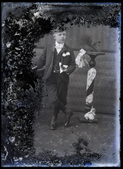 Staande foto van jongeman in feestkledij met wit hemd en vlinderdas, bloem op de boord, boek in de linkerhand, tgv Pl.Communie, Melle , 1910-1920