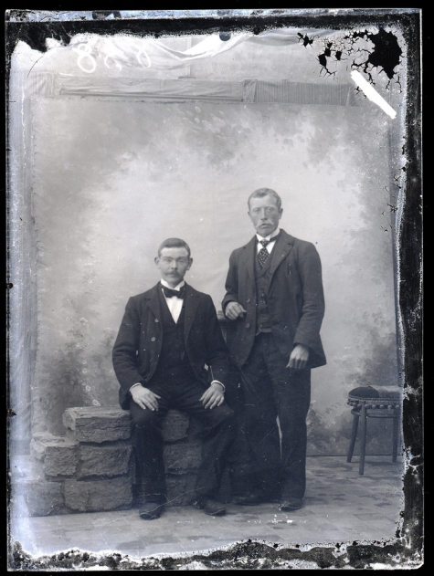 Portret van 1 staande en 1 zittende man in feestkledij zijnde kostuum en wit hemd met stropdas en vlinderdas, Melle , 1910-1920