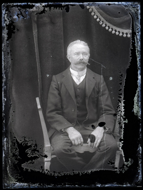 Zittend portret van man in feestkledij met kostuum en witte opstaande hemdskraag met stropdas, gemodelleerde snor en hoofdhaar met middenscheiding, Melle, 1910-1920