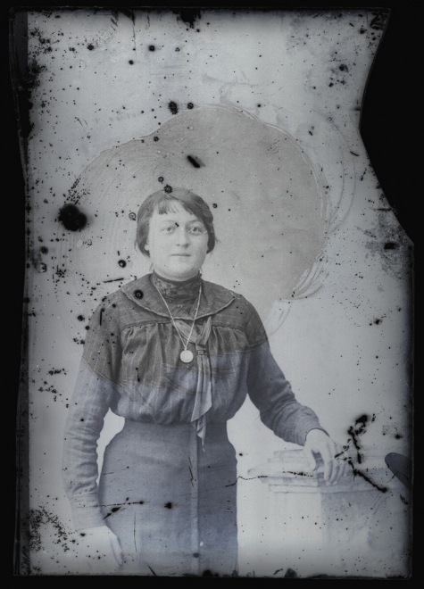 Staand portret van jonge vrouw in feestkledij bestaande uit lange rok en bloes met lange mouwen, stropdas en halsketting, bolvormig gekamd haar, Melle, 1910-1920