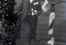 Staande foto van jongeman in feestkledij met wit hemd en vlinderdas, bloem op de boord, boek in de linkerhand, tgv Pl.Communie, Melle , 1910-1920