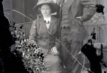 Portret van koppel in feestkledij, zittende vrouw met lange rok en vest, versierde bloeskraag, lange halsketting en opgespelde bloem en bloemversierde hoed, rechtstaande man in kostuum met hemd en stropdas, hoofddeksel, Melle, 1910-1920