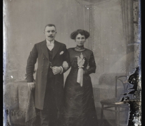 Staand studiofoto, koppel in feestkledij, man met lange jas en witte vlinderdas, vrouw met halsketting en bloementuiltje in de hand, Melle , 1910-1920