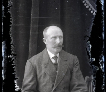 Zittend portret van man in feestkledij met witte hemdsboord en donkere stropdas, gemodelleerde snor en kalend voorhoofd, Melle, 1910-1920
